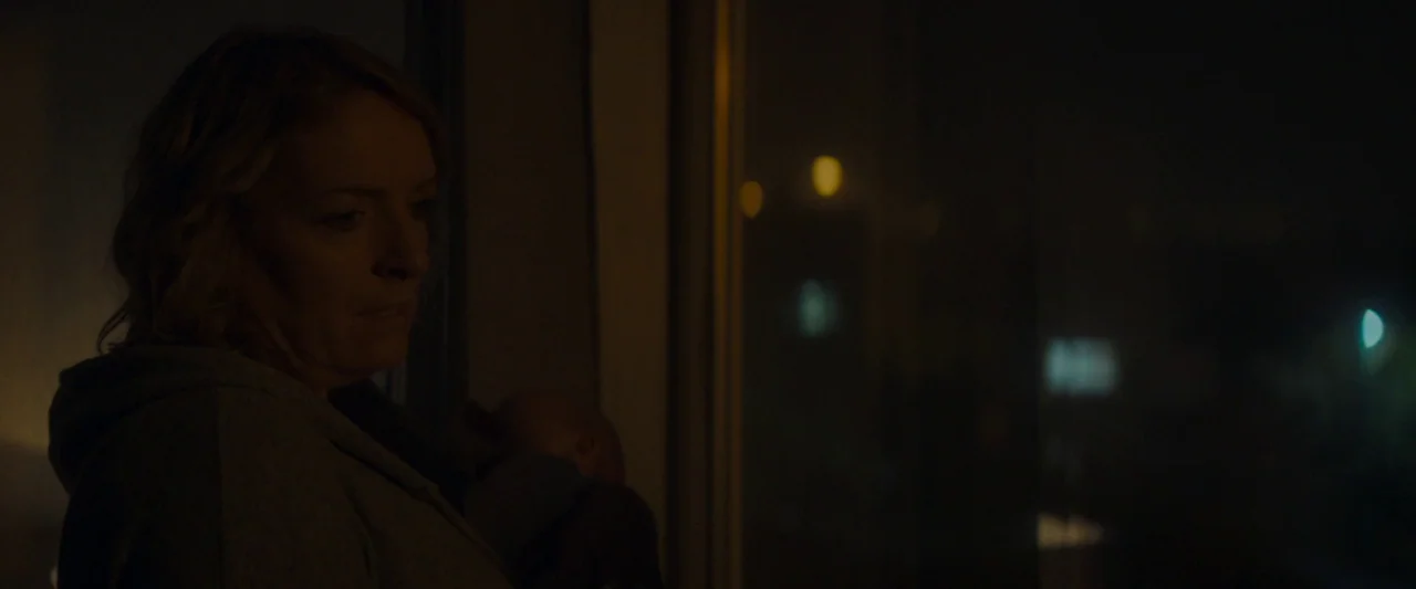 Ali & Ava (2021) movie still: medium — A dimly lit room interior at night, viewed through a window frame with city lights. A woman holding an infant…