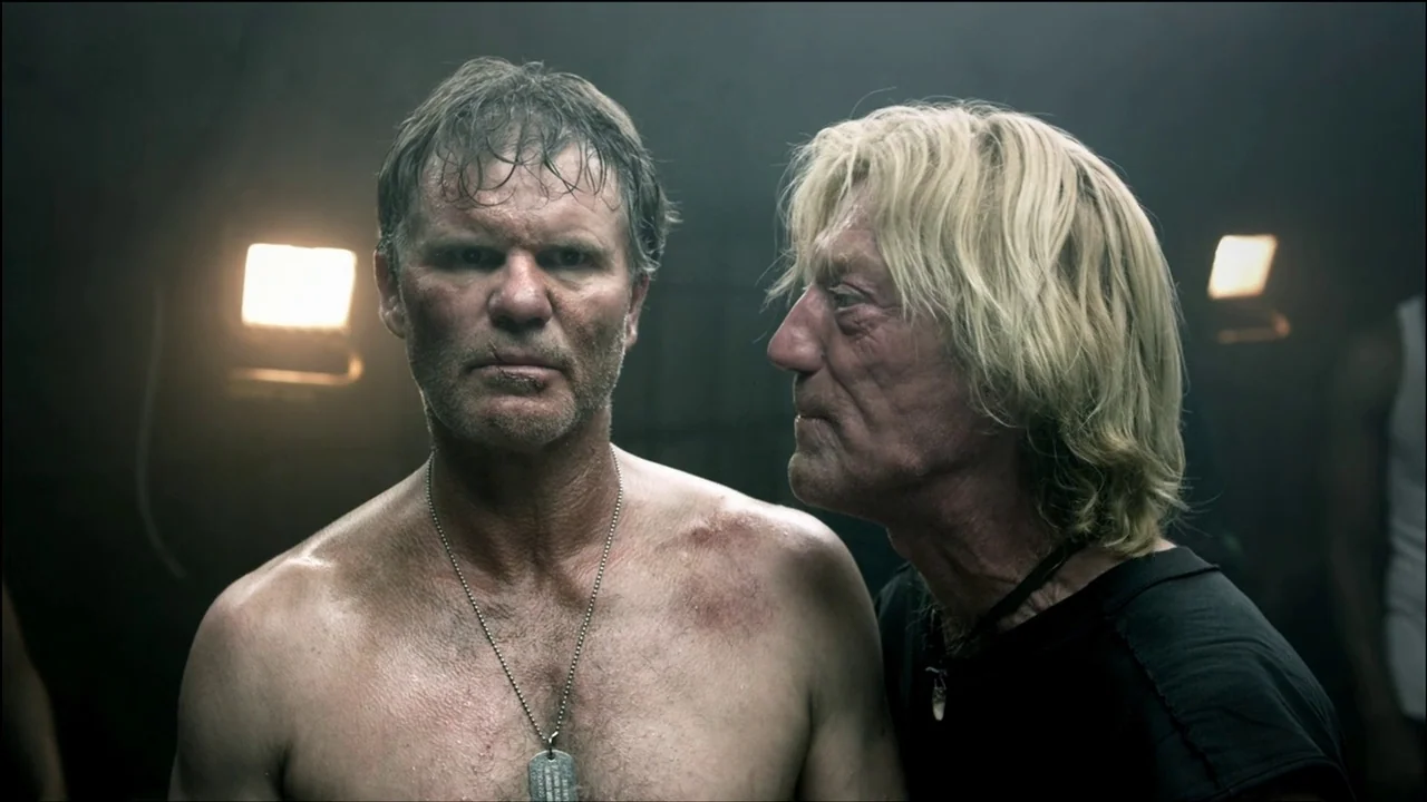 The ABCs of Death (2012) movie still: medium — Two men stand closely together in a dim, industrial environment filled with haze. One shirtless man…