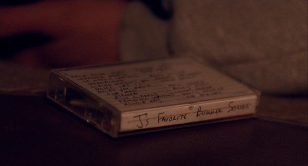 Adventureland (2009) movie still: closeup — A plastic cassette tape case rests on a dark surface in a dimly lit room. The white paper insert inside…