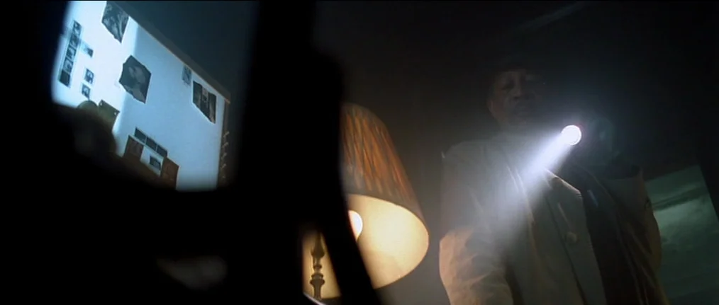 Se7en (1995) movie still: medium — A dark apartment interior contains a wall covered with pinned photographs and papers. An older man in a light tan…