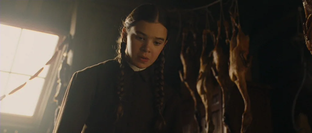 True Grit (2010) movie still: medium — A dark wooden larder features rows of hanging dressed poultry in the background. A young girl with braided…