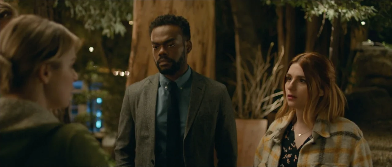 We Broke Up (2021) movie still: medium — A nighttime outdoor patio setting features large tree trunks and sparse greenery in the background. Three…