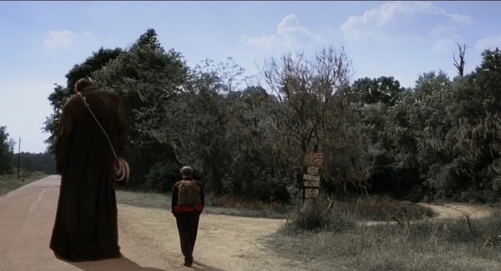 Big Fish (2003) movie still: wide — A rural road stretches past dense woods filled with trees draped in moss. Two men walk away from the viewer…
