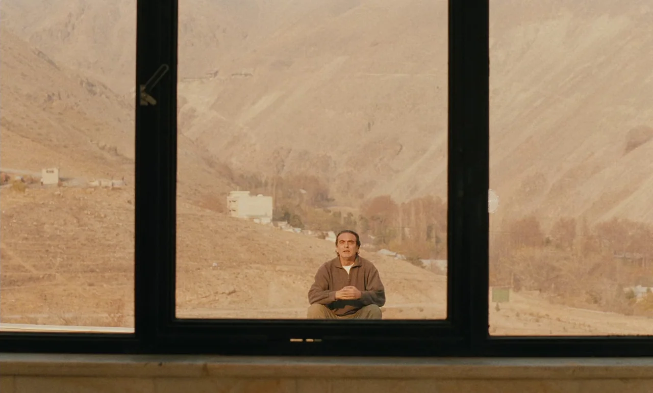 Taste of Cherry (1997) movie still: medium — An arid, rocky mountain range stretches across the horizon under a clear sky. A man sits alone on the…
