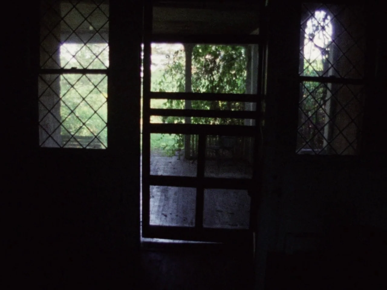 Grey Gardens (1975) movie still: establishing — An interior threshold looks out onto a shaded porch with lush greenery visible beyond. The doorway…
