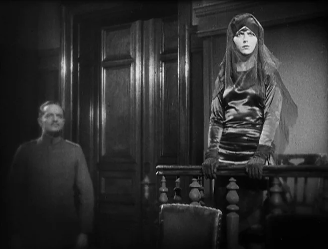 Pandora’s Box (1929) movie still: medium — A wood-paneled room with large double doors and a carved wooden railing. A woman in a veil leans on the…