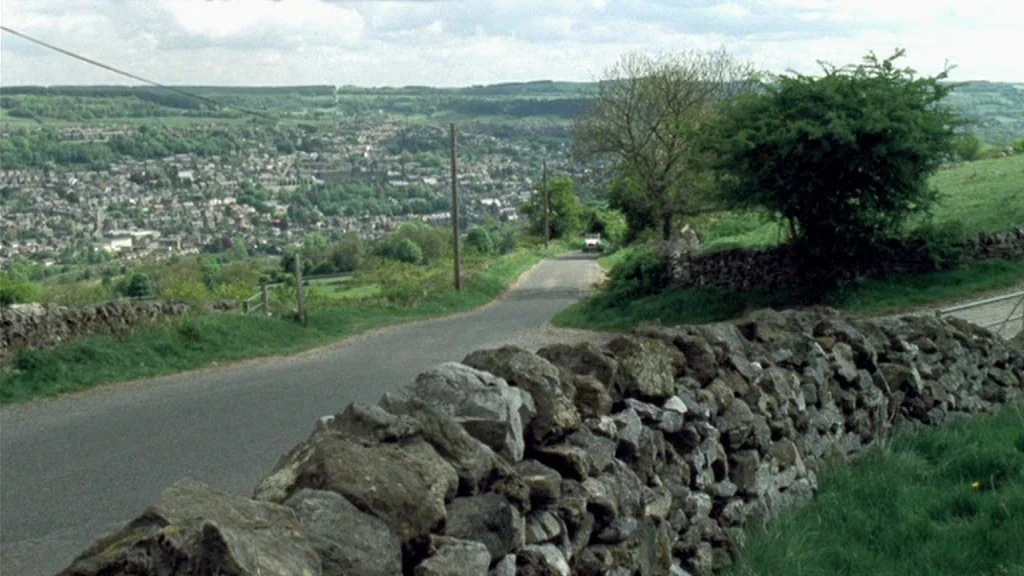 Dead Man’s Shoes (2004) movie still: establishing — A narrow country road winds downhill towards a distant town nestled in the valley. Dry stone…