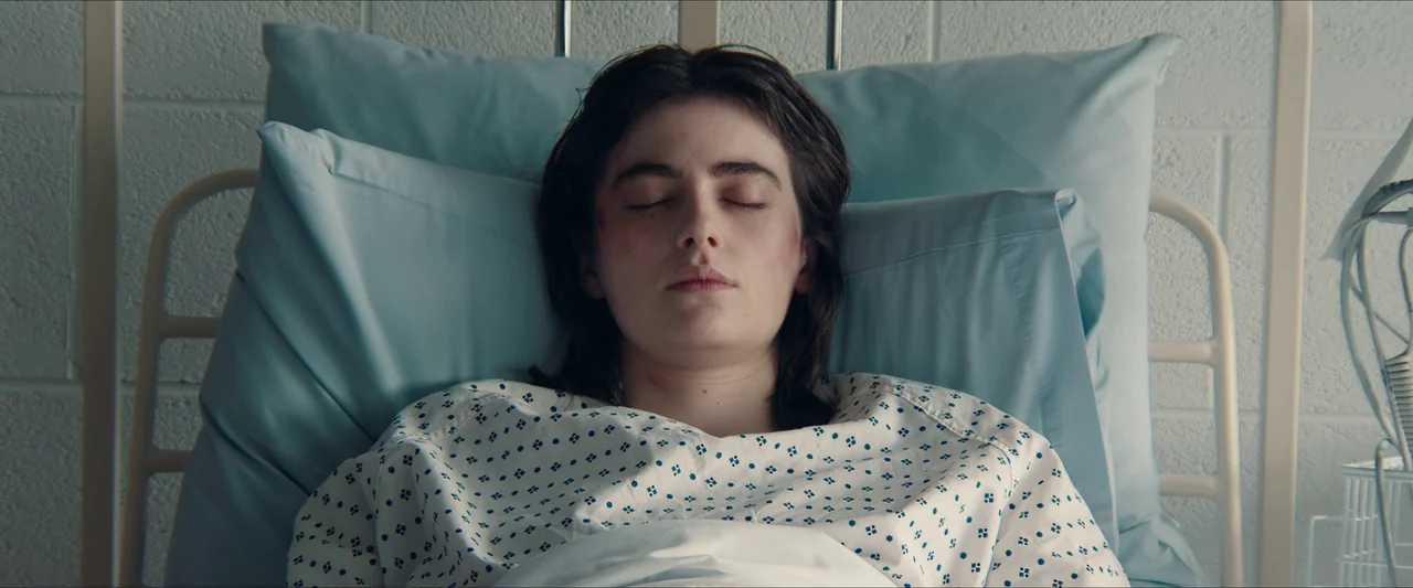 Double Blind (2023) movie still: medium — A person lies with eyes closed on a bed against a white cinder block wall. The patient rests on two light…