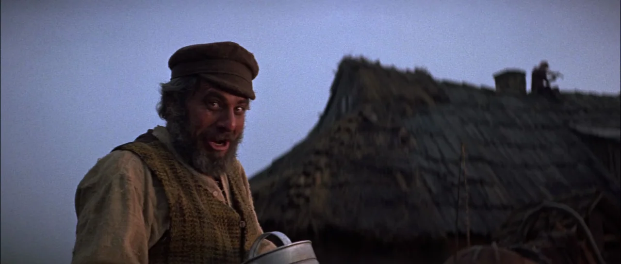 Fiddler On The Roof (1971) movie still: medium — An outdoor rural setting during dusk features a thatched roof building in the background. A bearded…