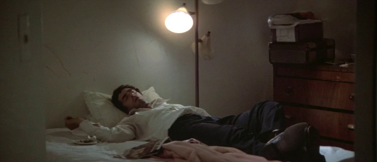 The Long Goodbye (1973) movie still: medium — A sparsely furnished bedroom with a plain bed and a wooden dresser. A man in formal wear rests on the…