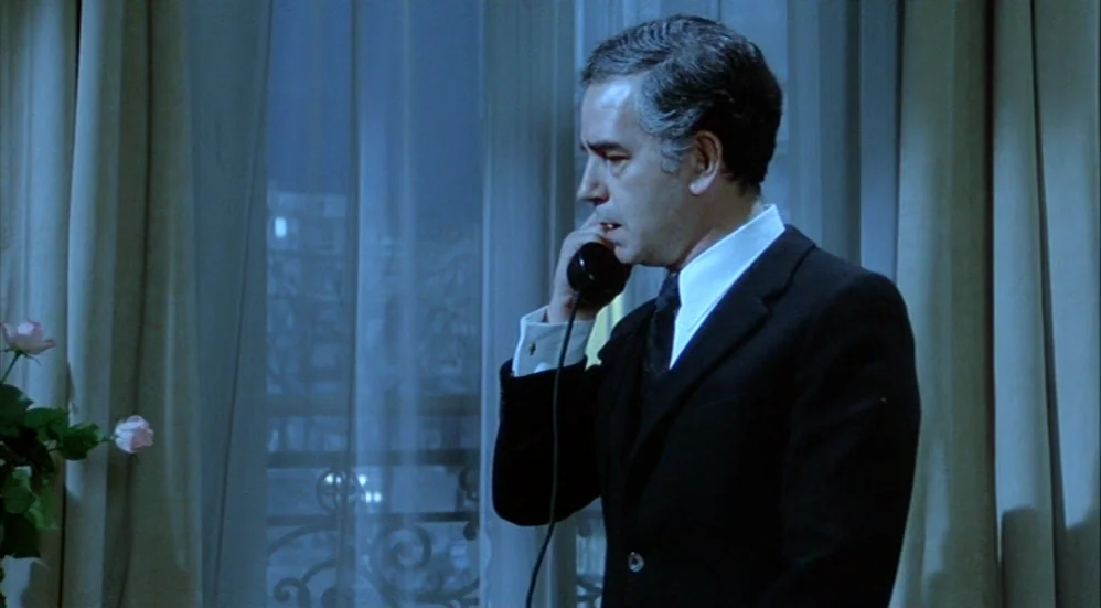 Un Flic (1972) movie still: medium — A hotel room with sheer curtains draped before a window overlooking a dark exterior. A man in a suit stands…