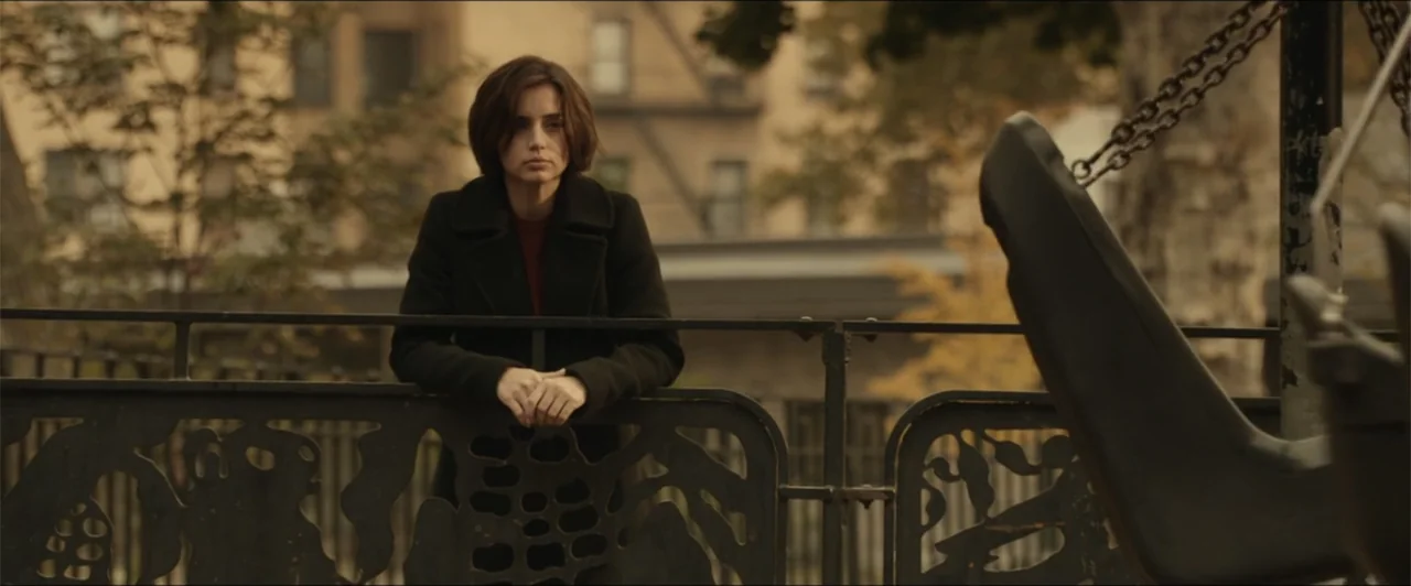 Exposed (2016) movie still: medium — A woman leans against an ornamental iron fence at a city playground. She stands still while facing forward…