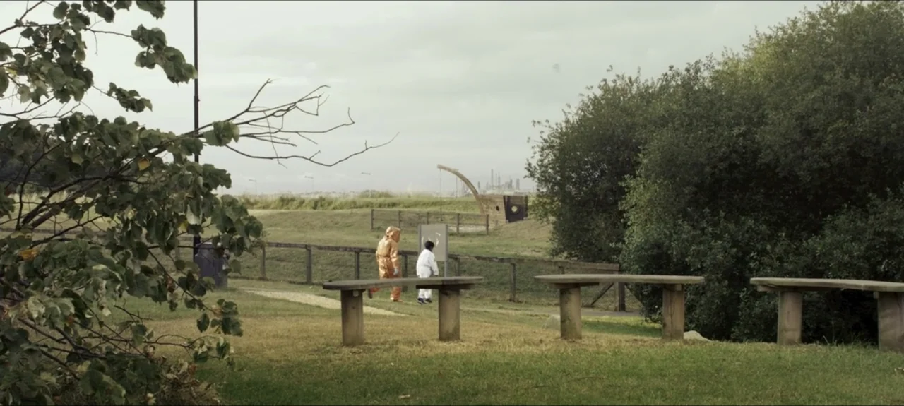 Containment (2015) movie still: wide — A grassy public park with wooden benches and a wire fence in the distance. Two figures in contrasting hazmat…