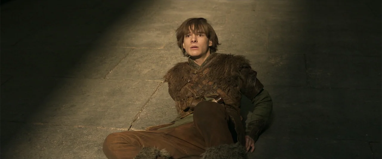How to Train Your Dragon (2025) movie still: medium — A dark stone floor interior is partially lit by a single high window. A young man sits on the…
