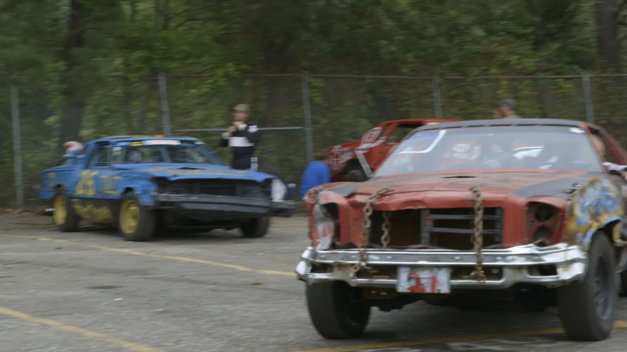 The Last Race (2022) movie still: medium — An asphalt track area shows damaged racing cars moving past a chain-link fence. Two men stand near their…