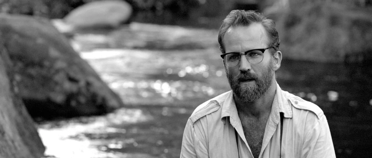 Embrace Of The Serpent (2015) movie still: medium — A man stands near a shallow river with smooth rocks in the background. The man looks toward the…