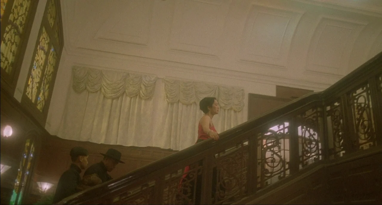Shanghai Triad (1995) movie still: wide — A grand interior staircase with ornate dark wood railings and stained glass accents. A woman in a red…