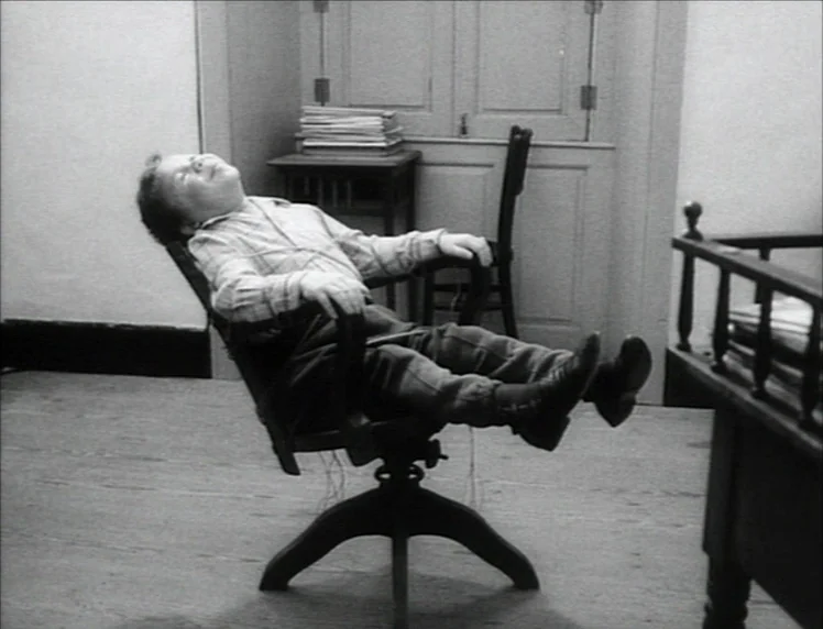 Even Dwarfs Started Small (1970) movie still: fullbody — A sparsely furnished room contains a vintage swiveling chair and a wooden desk. A boy sits…