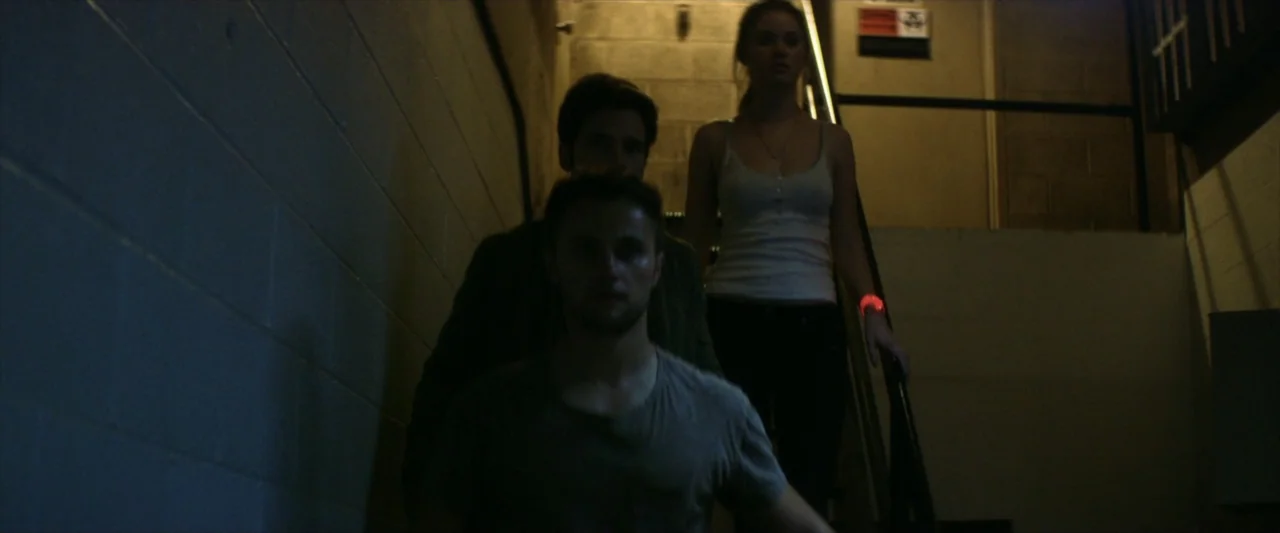 Tell Me How I Die (2016) movie still: medium — A dark concrete stairwell with cinder block walls and a metal handrail. Three young adults walk down…