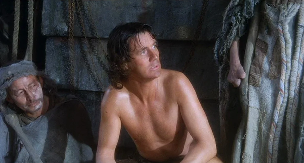 Life of Brian (1979) movie still: medium — A dimly lit dungeon space features large stone blocks and hanging iron chains. A shirtless man sits…