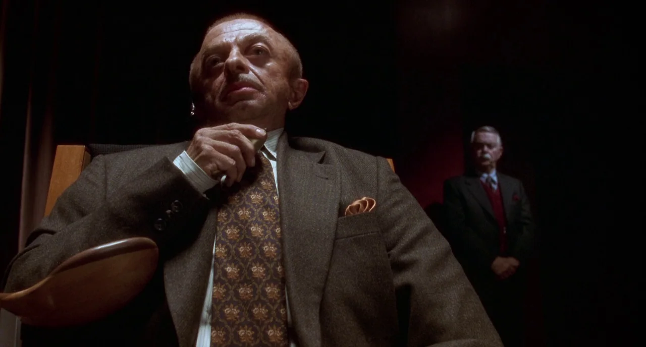 Mulholland Drive (2001) movie still: medium — A dimly lit room features a foreground subject sitting in a wooden-armed chair. An older man in a suit…