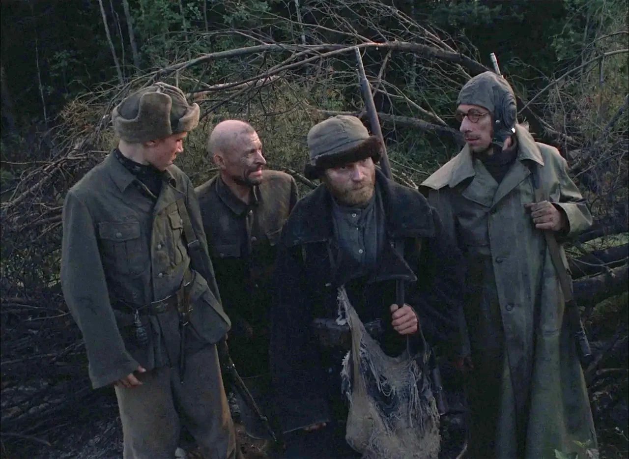 Come and See (1985) movie still: medium — A group of soldiers stand in a dark, brush-filled forest during the dim twilight hours. The men are…