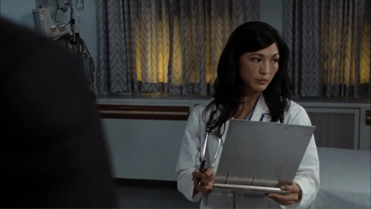 Driven to Kill (2009) movie still: medium — A sterile medical room features patterned curtains and a medical monitor in the background. A doctor…