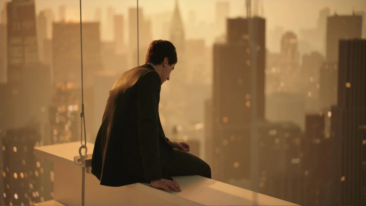 Megalopolis (2024) movie still: medium — A man sits on the high edge of a skyscraper with a dense city skyline behind. He gazes downward at the…