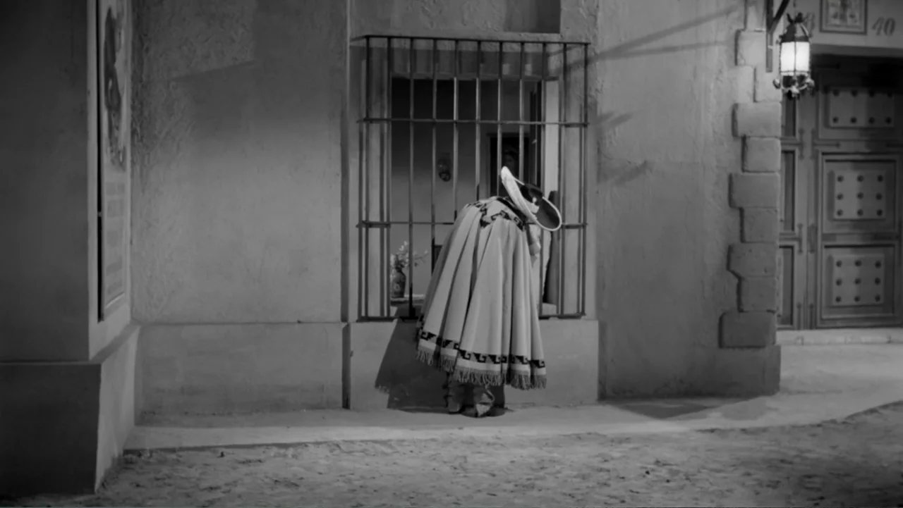Escuela para solteras (1965) movie still: medium — A dark city street features an adobe-style wall with a single barred window. A man in a heavy…