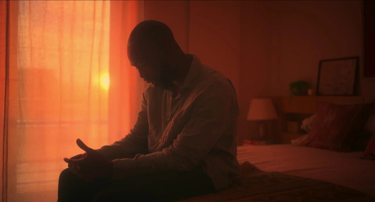 Men (2022) movie still: medium — A man sits on the edge of a mattress in a dimly lit space. The intense glow of a sunset streams through orange…