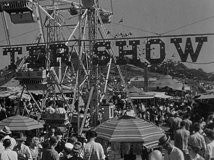 Nightmare Alley (1947) movie still: wide — A crowded outdoor carnival grounds features a large Ferris wheel and striped umbrellas. Numerous…