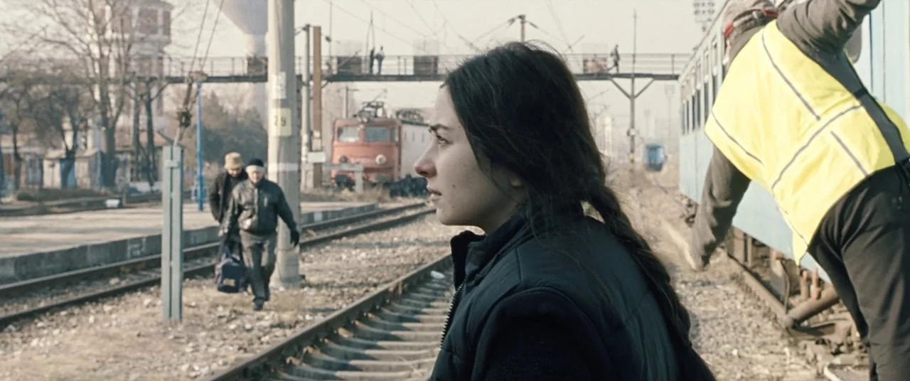 Beyond the Hills (2012) movie still: medium — A desolate railway yard with metal tracks and weathered infrastructure under an overcast sky. A young…