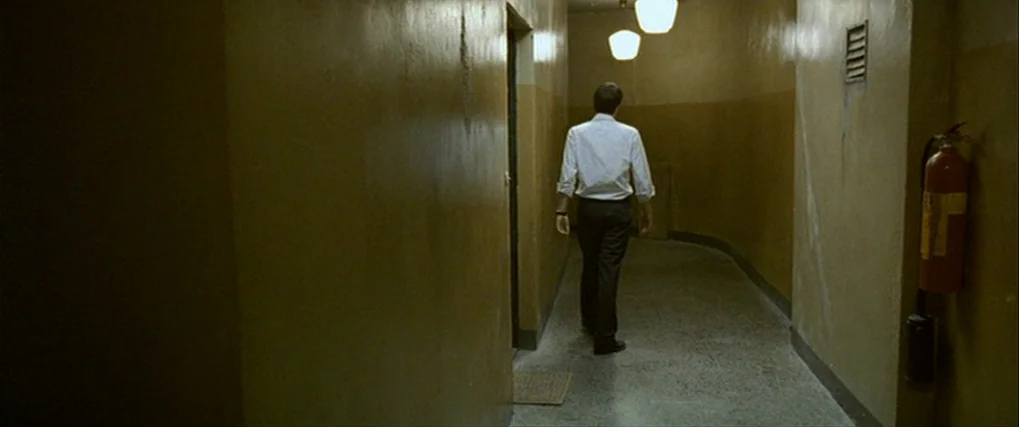 Next Door (2005) movie still: fullbody — A narrow, dimly lit hallway with plain ochre-painted walls and a grey tiled floor. A man walks away from…