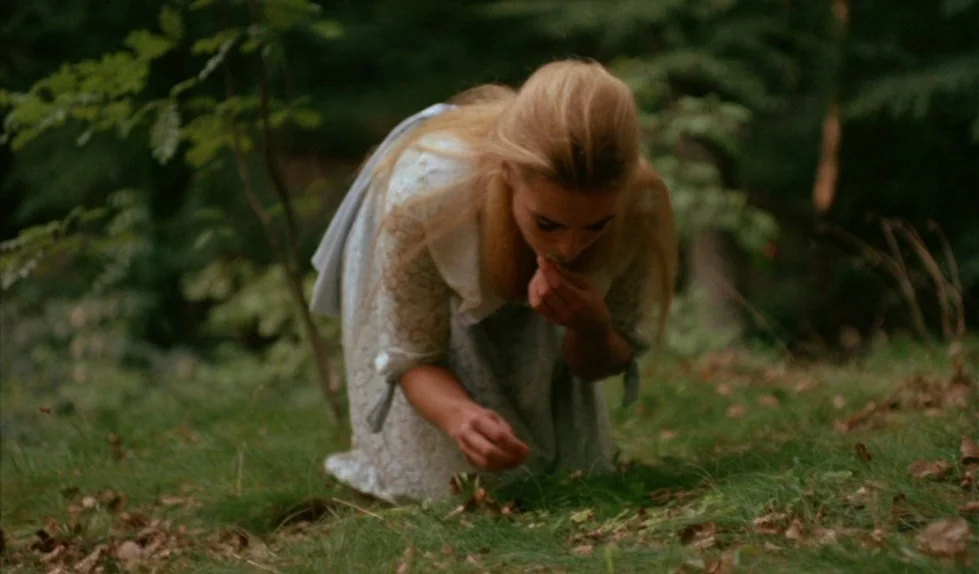 Elvira Madigan (1967) movie still: medium — A woman kneels in a lush woodland area with grass and fallen leaves. She leans forward to examine or…
