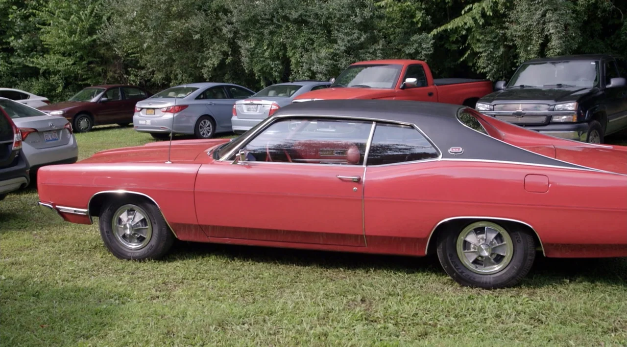 The Divided Soul of America (2019) movie still: medium — A classic red coupe with a black vinyl roof sits on a grassy field. Several modern sedans…