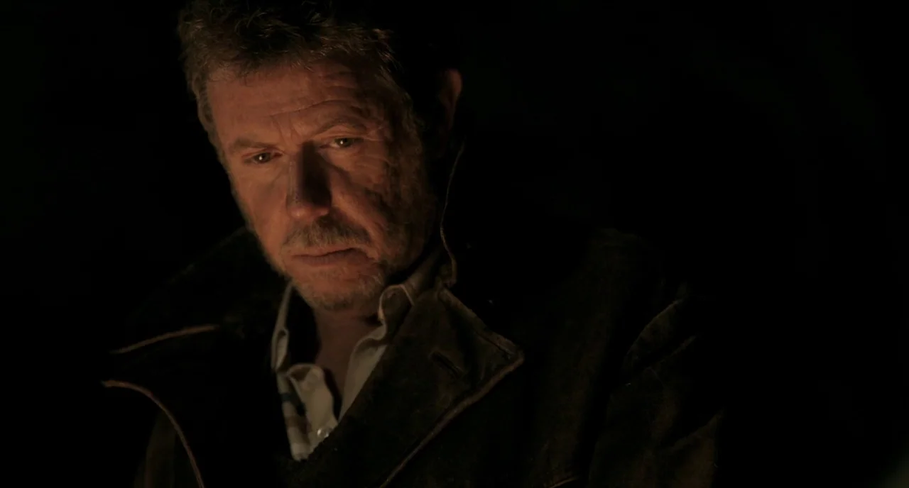 The Selfish Giant (2013) movie still: closeup — A dark, indistinct exterior area is illuminated only by a nearby, unseen fire. An older man wearing…