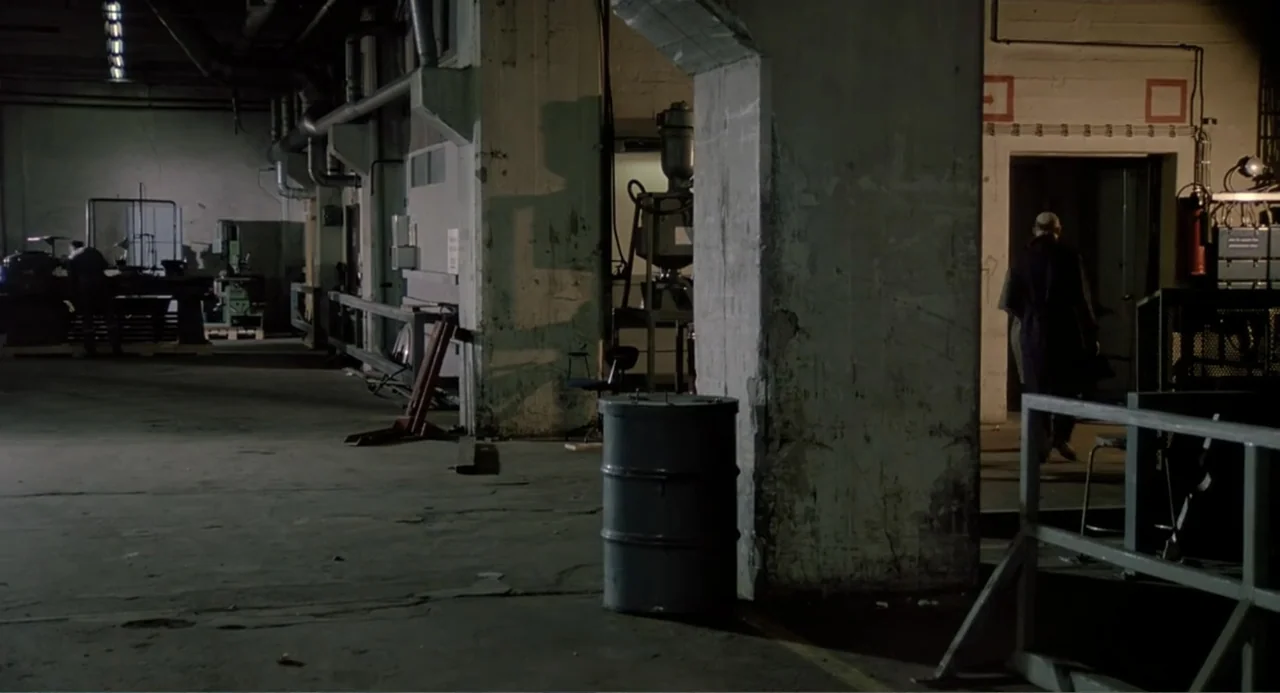 The Match Factory Girl (1990) movie still: wide — An expansive industrial warehouse features exposed concrete pillars and heavy machinery in the dim…
