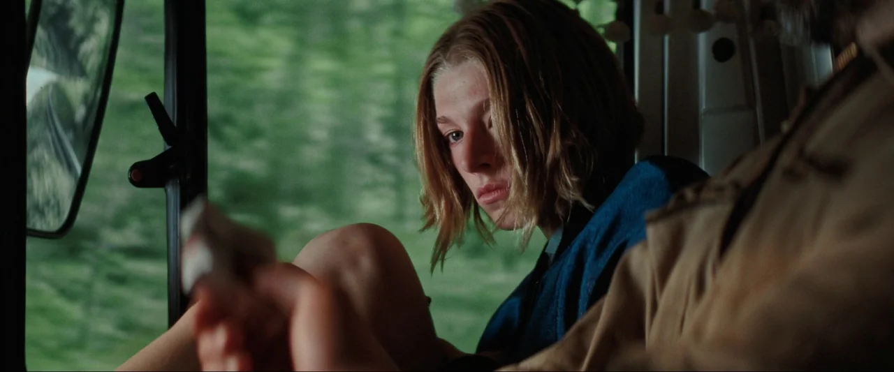 Cuckoo (2024) movie still: medium — Inside a moving van, a woman with short hair sits with her knees pulled up. Green vegetation is visible through…