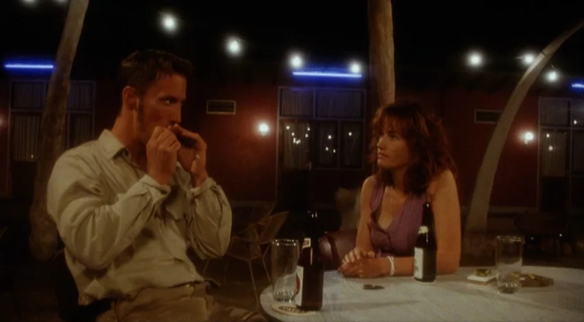 Dust Devil (1992) movie still: medium — A night scene on an outdoor patio with reddish walls and string lights above. A man lights a cigarette while…