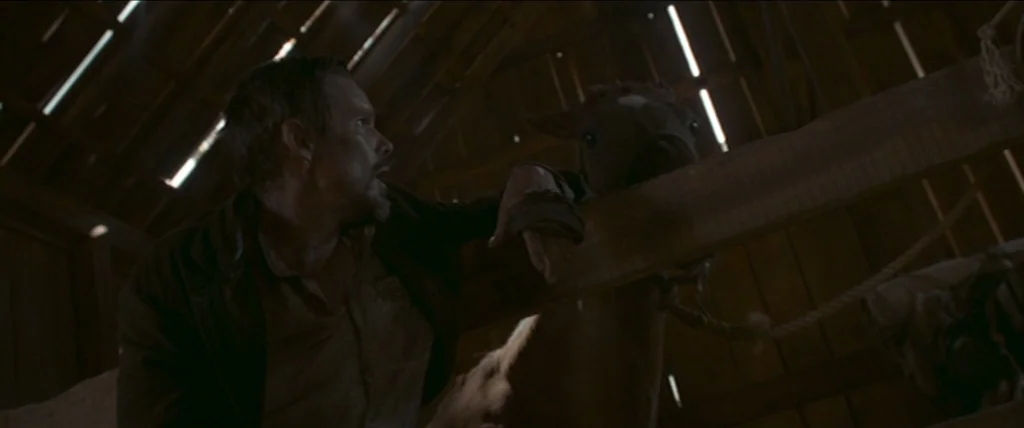In A Valley of Violence (2016) movie still: medium — A dark wooden structure has gaps in the roof allowing light to stream through. A man leans…