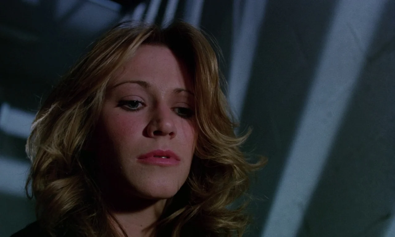 Rabid (1977) movie still: closeup — A dark, dimly lit corridor with angled shadows cast against a wall. A young woman with blonde hair looks down at…