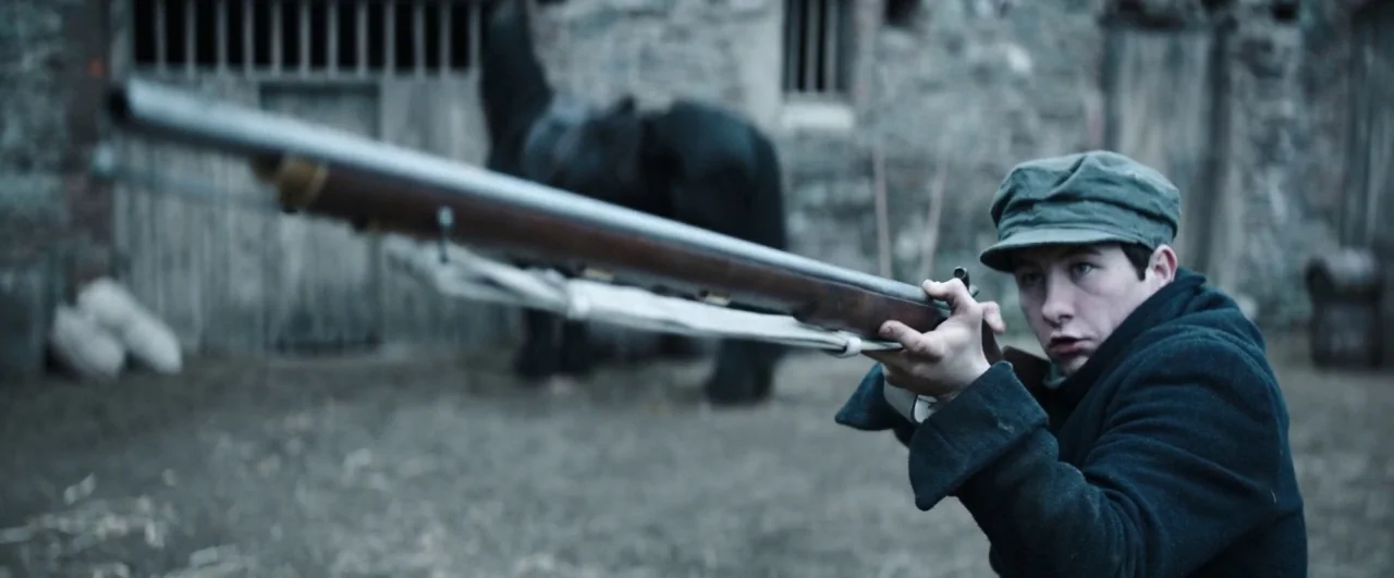 Black ’47 (2018) movie still: medium — A rural courtyard features a weathered stone wall and wooden barn doors. A young man aims a long,…