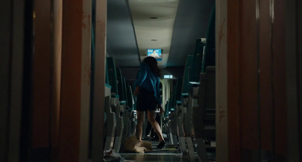 Train To Busan movie still: wide — The interior of a commuter train car features rows of teal-colored seats and overhead monitors. A woman stands in…