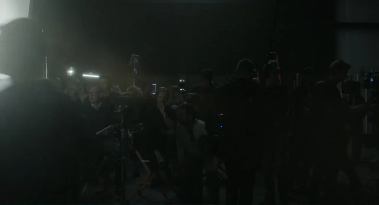 May December (2023) movie still: wide — A dimly lit production studio contains numerous crew members sitting in folding chairs. The background…