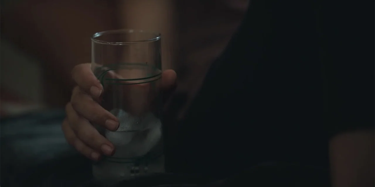 Equal Standard (2021) movie still: closeup — An interior space is dimly lit and largely obscured by shadows. A person holds a glass filled with…