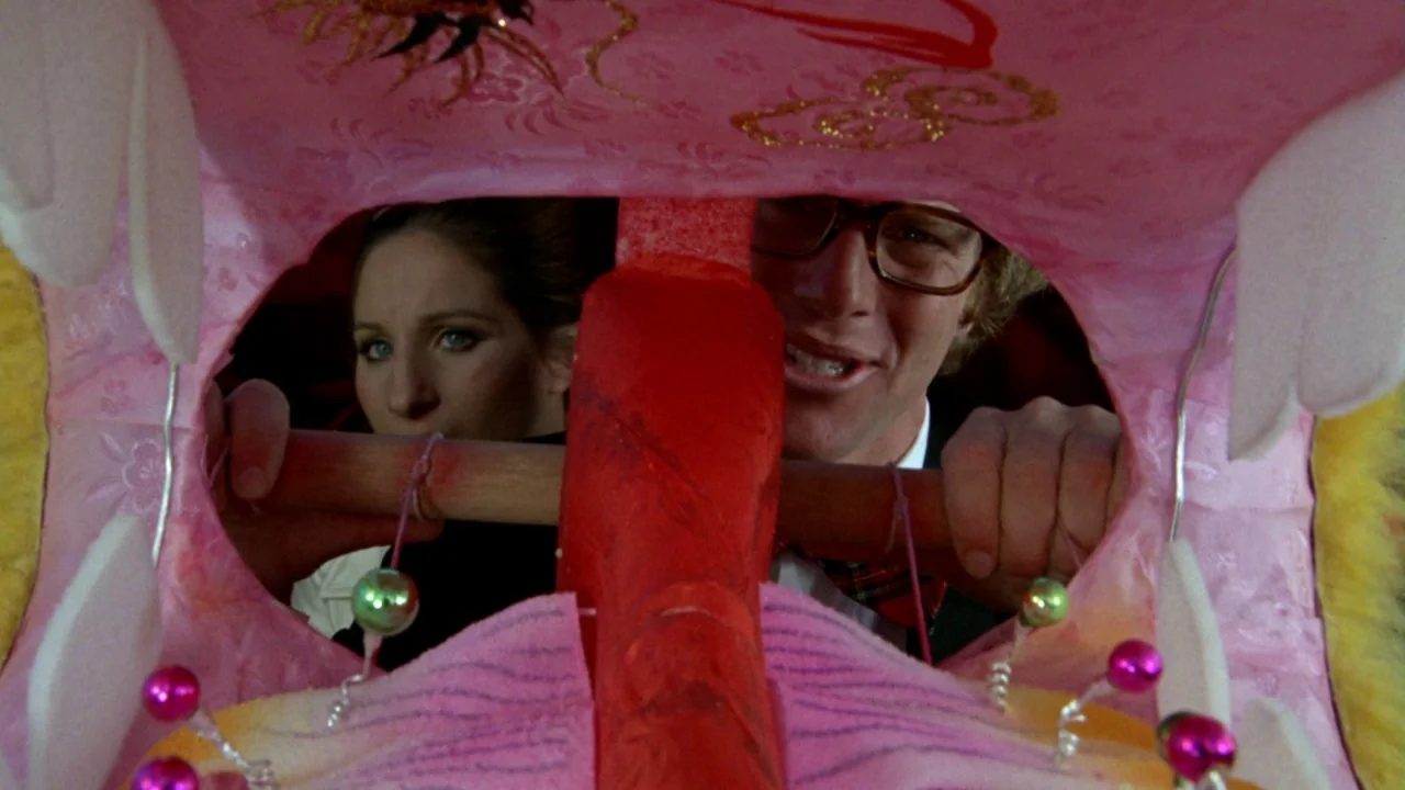 What’s Up, Doc? (1972) movie still: medium — A man and a woman look through the mouth of a large pink prop. The prop is decorated with glitter and…