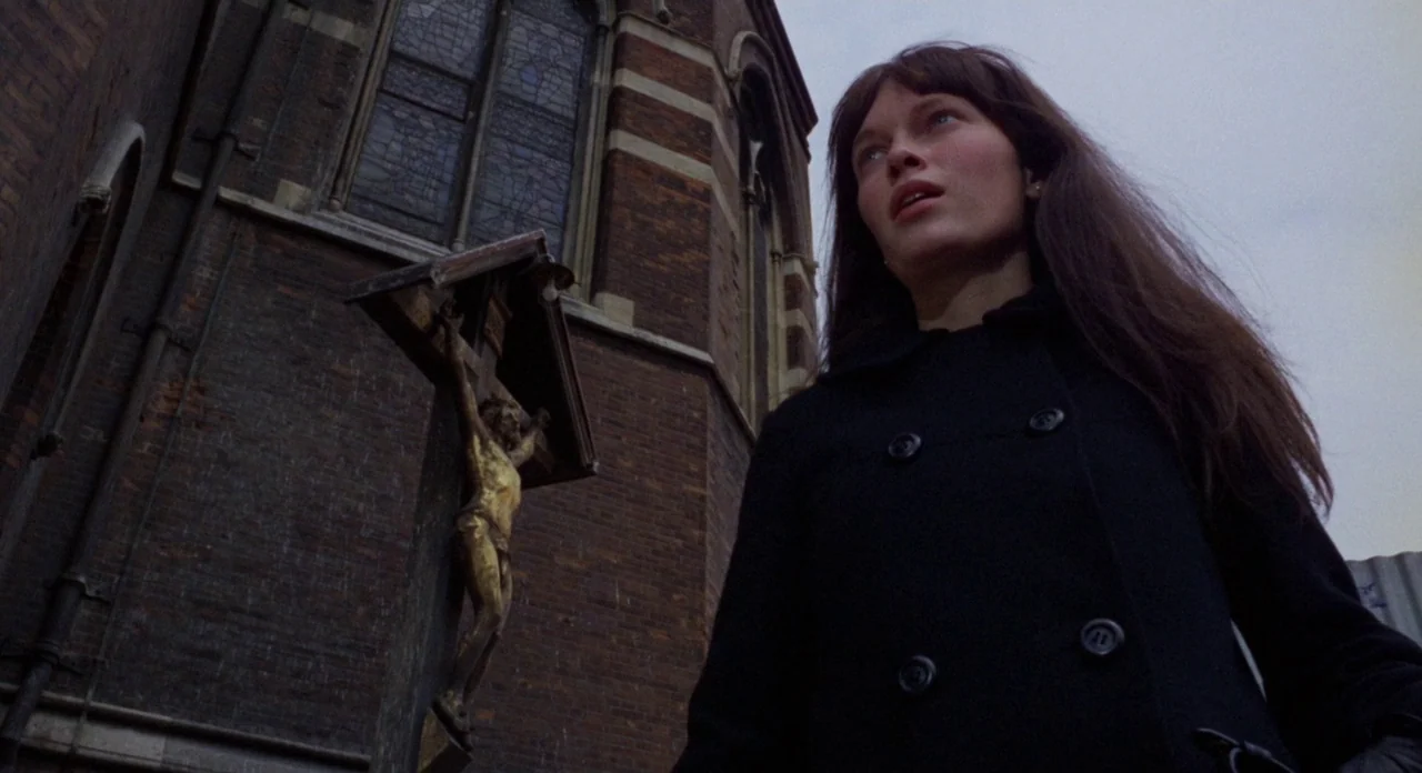 Secret Ceremony (1968) movie still: medium — A red brick wall of a church structure features a large stained glass window. A woman in a dark coat…