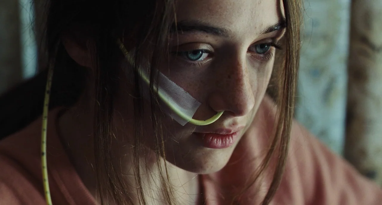 The Killing of a Sacred Deer (2017) movie still: closeup — An interior space with blurred, patterned wallpaper in the background. A young woman with…