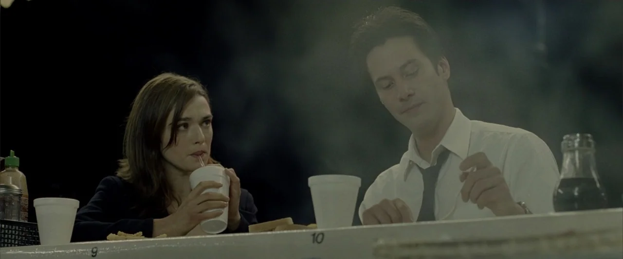Constantine (2005) movie still: medium — A dark, outdoor dining counter is dimly lit with white cups and plates present. A man and a woman sit at…