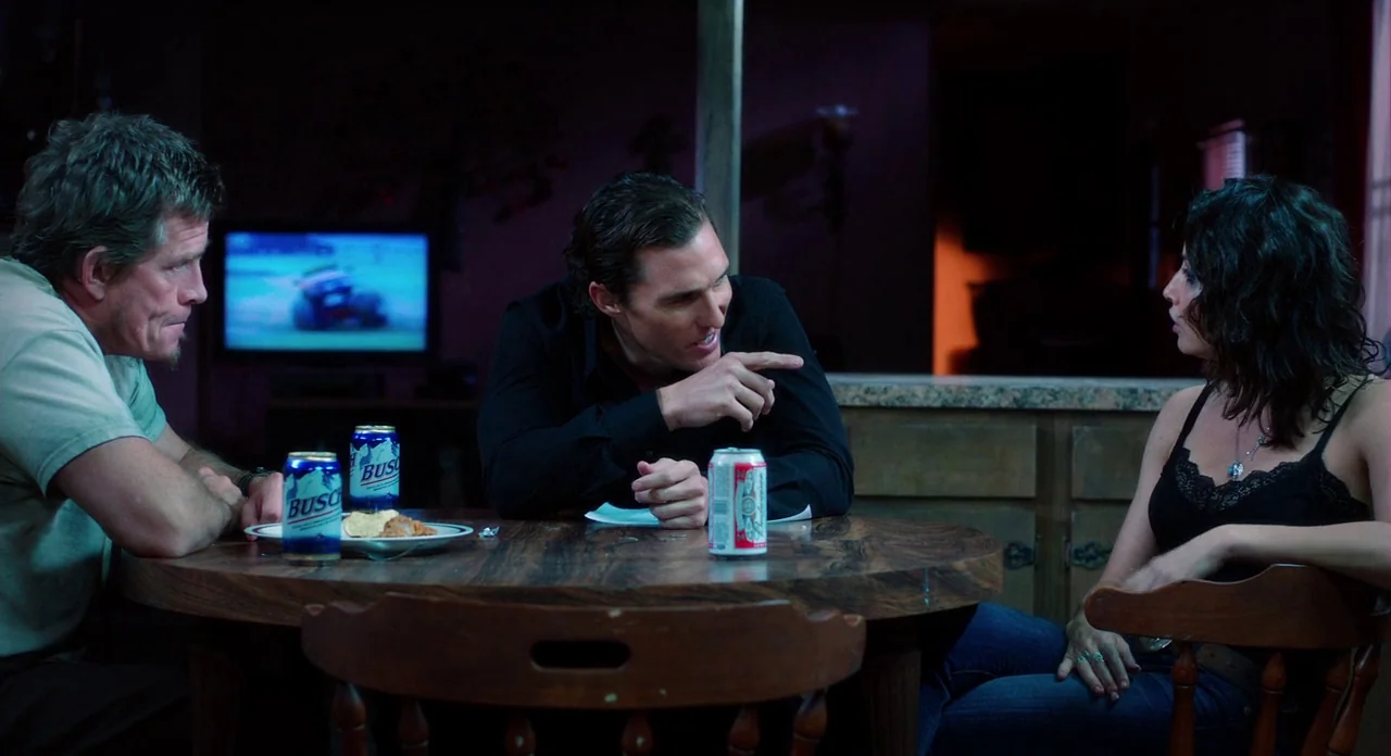 Killer Joe (2011) movie still: medium — A dark dining room with a round wooden table, blurred television in the background. Two men and a woman sit…
